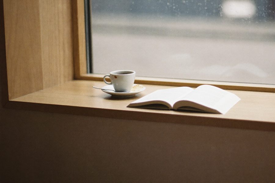Den brede vindueskarm indefra, med en kop og en opslået bog og regn på ruden.